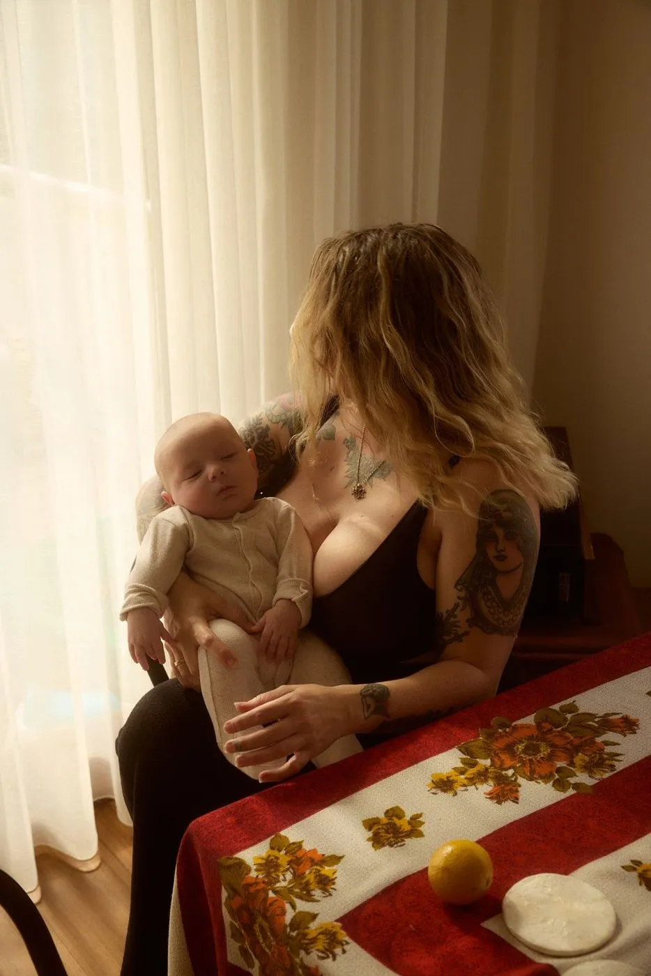 A mother holds her sleeping baby by a sunlit curtained window
