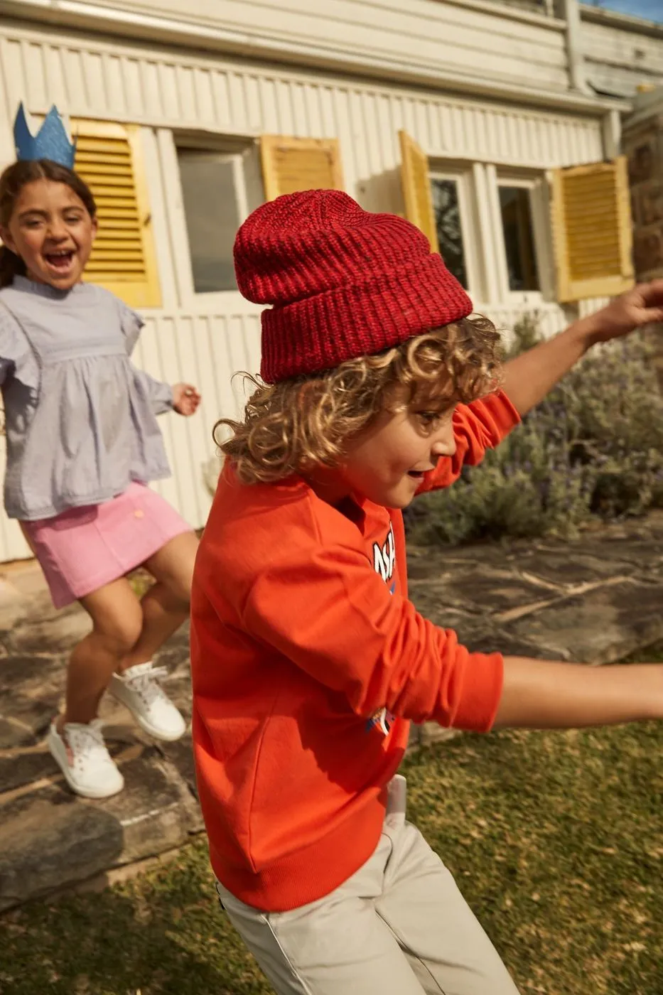 A boy in a red beanie and a girl in a crown run laughing past a weatherboard house